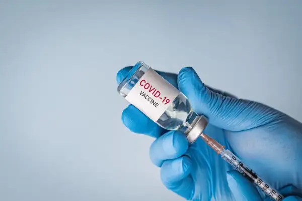 Ręka w niebieskiej rękawiczce trzyma fiolkę z napisem "COVID-19 VACCINE" i strzykawkę, gotowa do szczepienia.